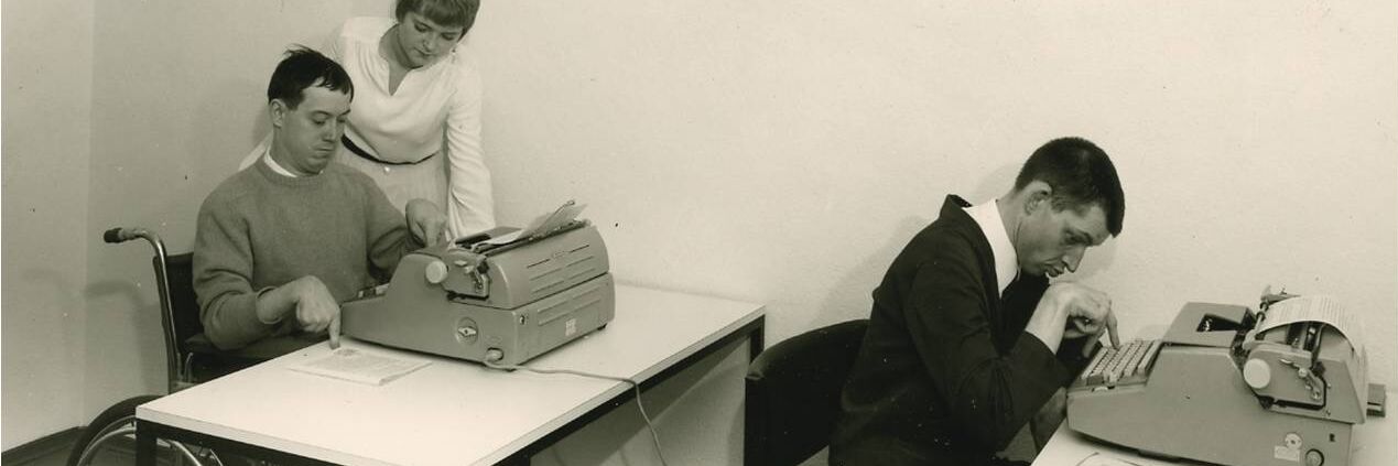 Das Bild zeigt zwei Männer an einem Schreibtisch die mit Schreibmaschinen schreiben.